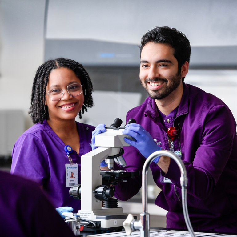 A smiling male and a smiling female laboratory technologist looking at a microscope