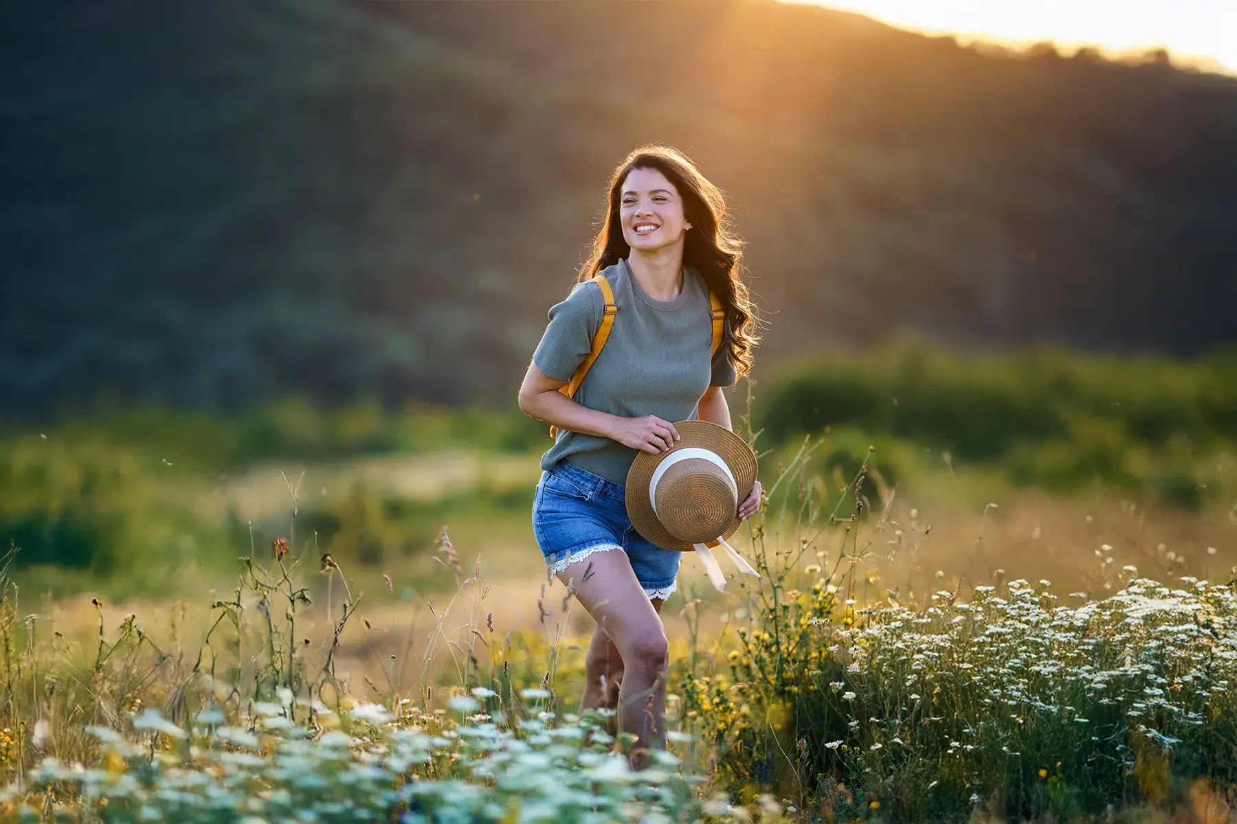 Happy woman is walking on the grass, holding a hat in her hand, while the sun sets behind the mountain.