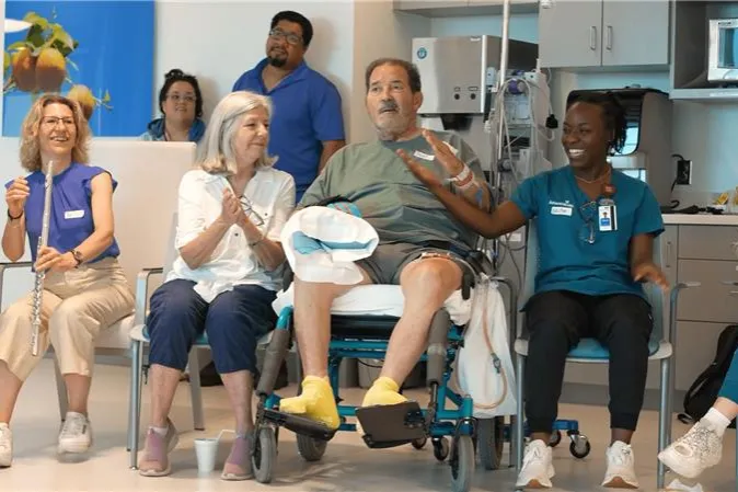 Patient seated in a wheelchair with family member and clinicians participating in a music activity in a hospital room.