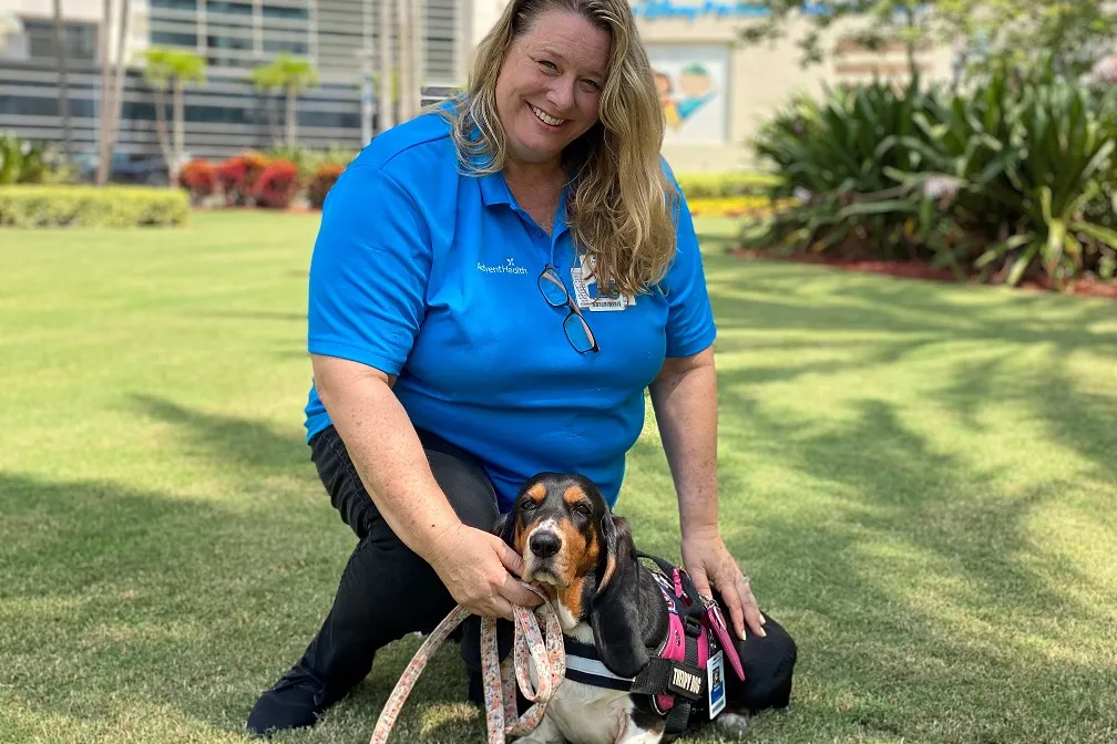Woman kneeling on grass with a therapy dog wearing a service harness.